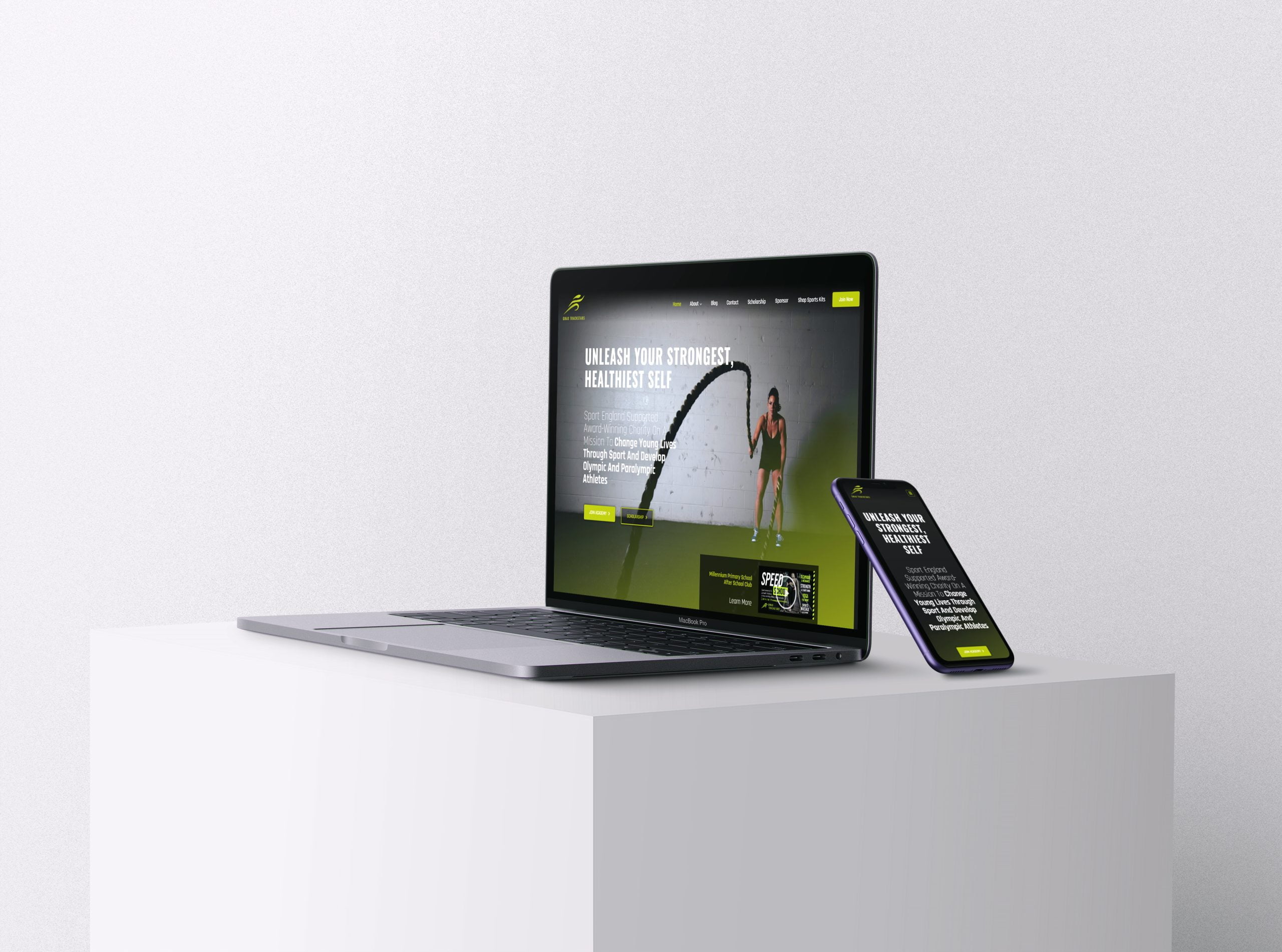 A laptop computer with an open lid sits on a desk next to a smartphone. The laptop screen displays a fitness website with the text "UNLEASH YOUR STRONGEST HEALTHIEST SELF" in large blue letters. The text is centered on the screen with a green gradient background. There is also a logo in the top left corner but it can't be discerned from the image.
